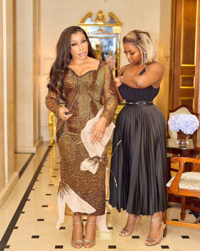 Rita Dominic stands in a grand hallway wearing a shimmering gold and blush TUBO gown, while Tubo makes final styling adjustments.